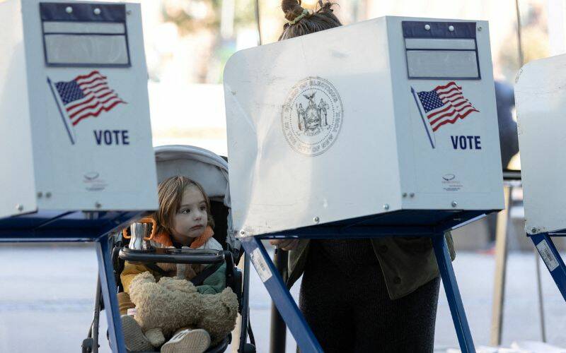 A girl in a stroller watches a woman cast a ballot. The woman is shielded by a privacy screen that reads vote with the American flag depicted on it.